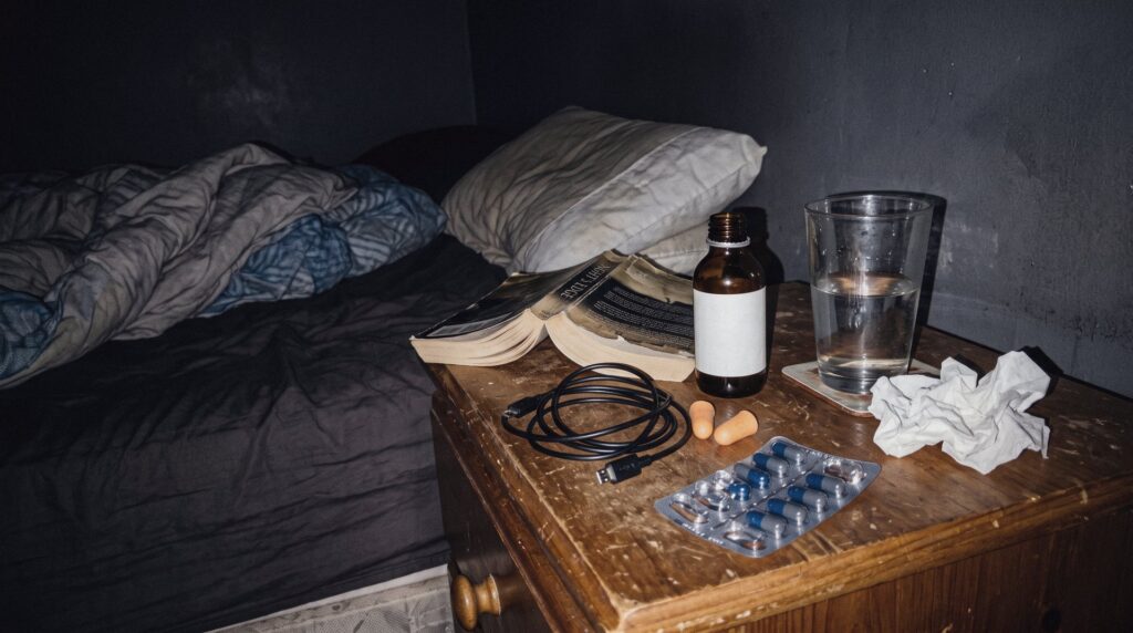 Mesilla de noche desordenada junto a una cama deshecha, con un frasquito de aceite, un vaso de agua, tapones para los oídos y un blíster de cápsulas