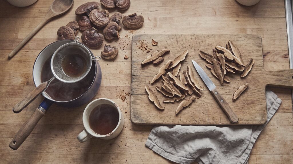 Láminas de reishi seco sobre una tabla de cortar con un cuchillo, junto a un cazo con infusión y una taza