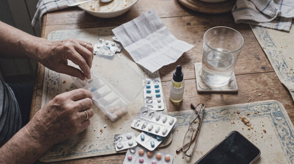 Manos de una persona mayor organizando pastillas en un pastillero semanal, con un frasco de CBD, blísteres de medicamentos, prospecto y un vaso de agua sobre la mesa