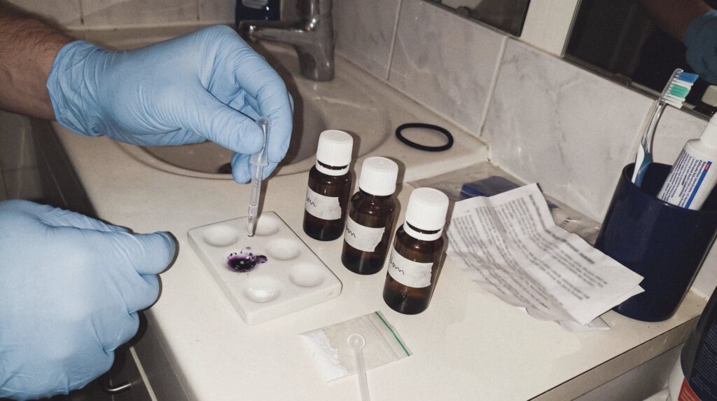 Manos con guantes de nitrilo aplicando una gota de reactivo en una placa de ensayo junto al lavabo, con varios frascos de reactivo